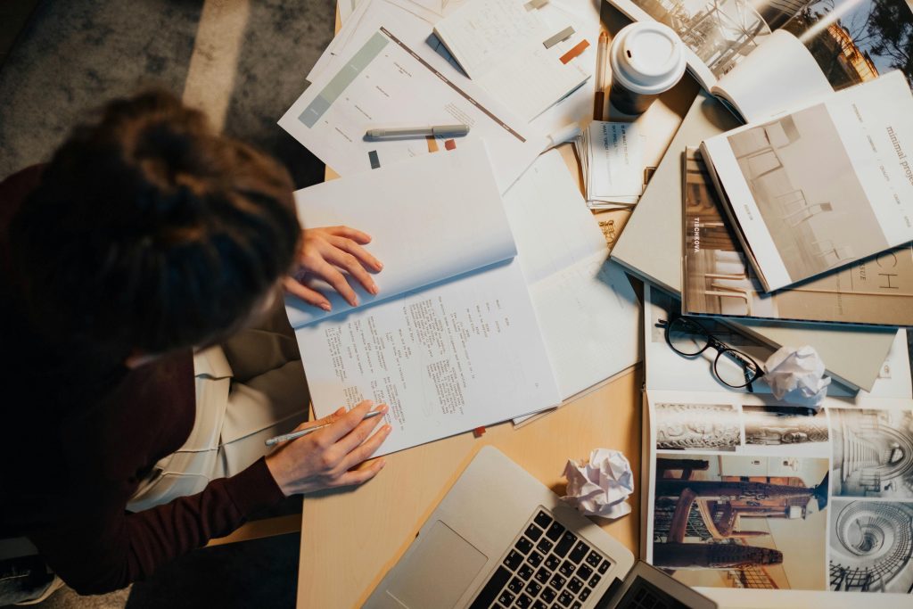 Overhead view of a cluttered work desk with a person writing notes, surrounded by papers, laptop, and coffee cup.